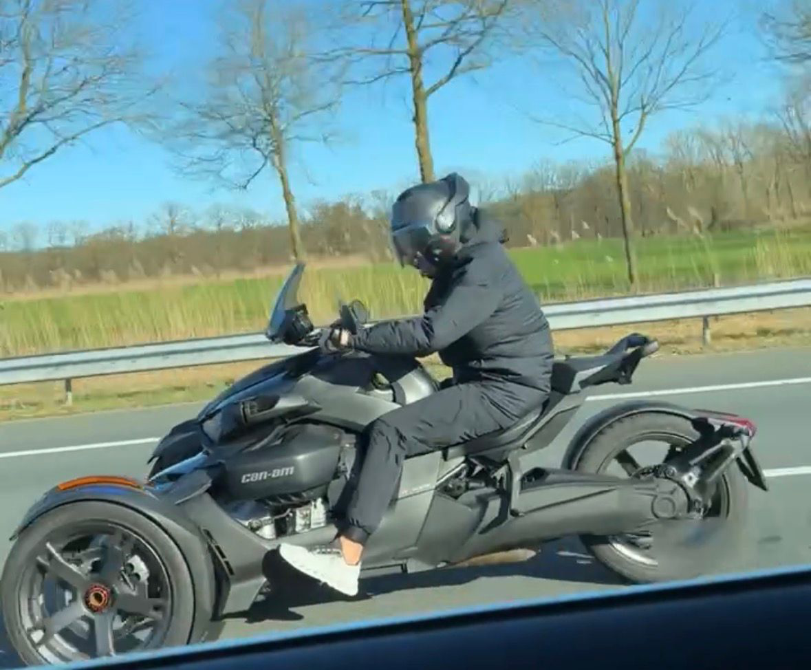 A man riding a Can-Am motorcycle on a highway, wearing a black helmet and jacket, with a grassy field and trees in the bac...