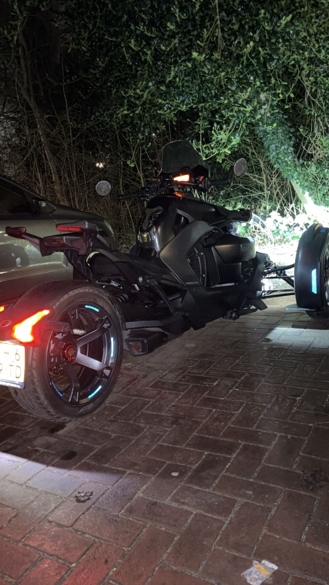 Can-Am Ryker bij nacht gezien van schuin achteren op een natte Amsterdamse oprit, met verlichte velgen en remlichten die reflecteren op de rode klinkers