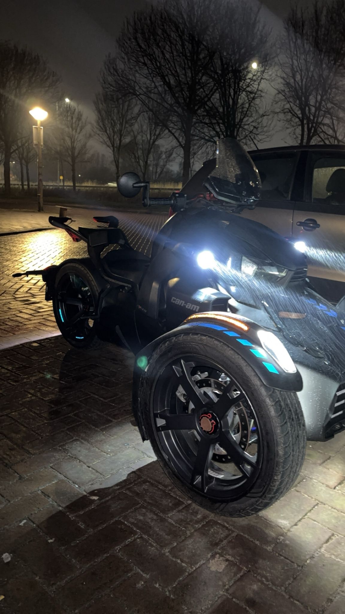 Can-Am Ryker bij nacht op een natte Amsterdamse straat, vlakbij geparkeerde auto’s en bomen, met felle koplampen en stadsverlichting die weerspiegelen op het wegdek