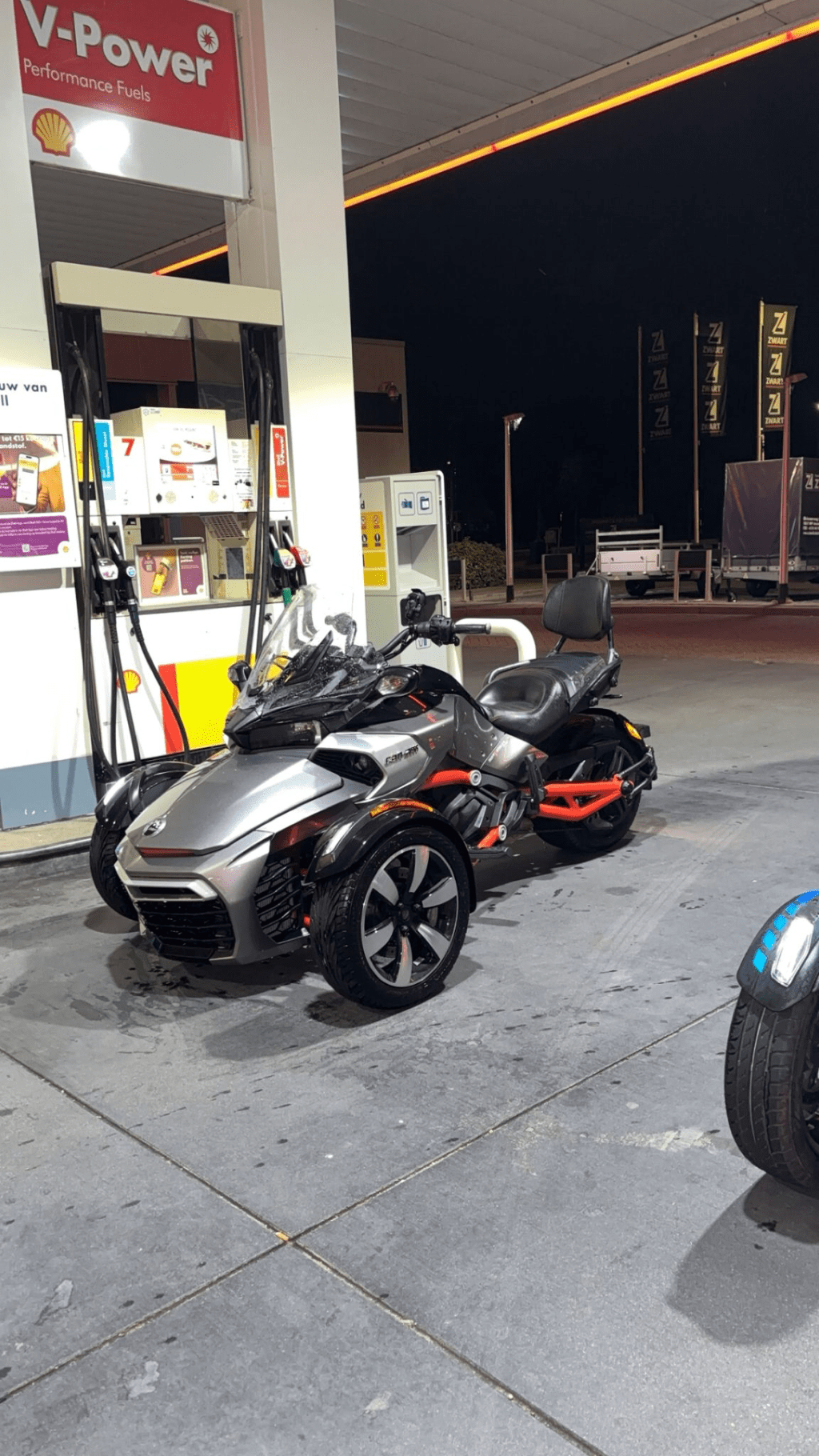 A silver and black Can-Am Spyder motorcycle is parked at a Shell gas station at night. The vehicle has a sleek design with...